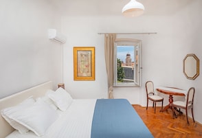 Quarto com cama, janela com vista para um edifício, mesa com cadeiras e ar-condicionado fixado na parede.