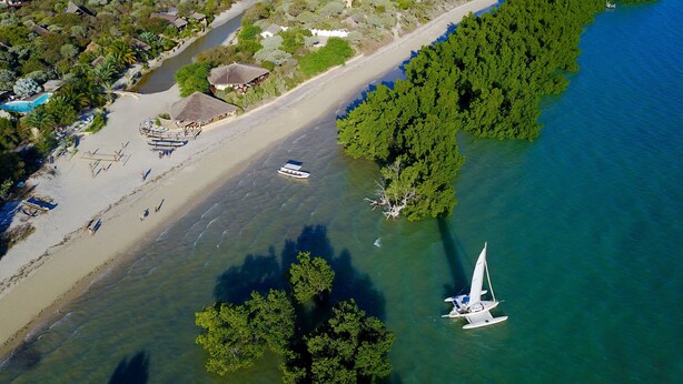 Vue aérienne d’une zone côtière avec une plage de sable, un petit bateau et un voilier ancré près du rivage.