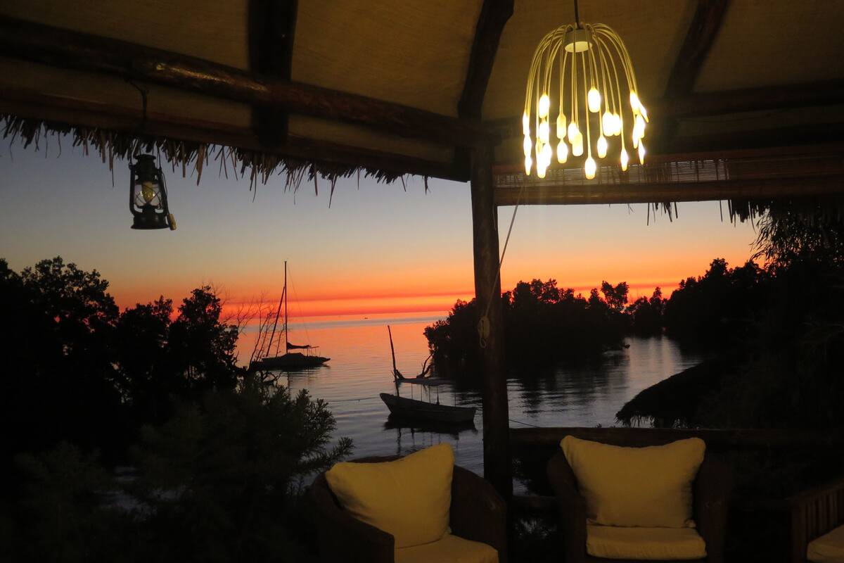 A room with a view of the sunset over water, featuring a hanging light fixture and two chairs.
