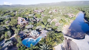 Vue aérienne d’un complexe hôtelier comprenant plusieurs bungalows aux toits de chaume, une piscine centrale et une plage.