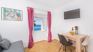 A room with a wooden floor, a grey sofa, a dining table with two chairs, a TV mounted on the wall, and a window with pink curtains. - Apartment View (Dubrovnik)