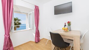 A room with a wooden floor, a table with chairs, a TV mounted on the wall, and a window with pink curtains. - Apartment View (Dubrovnik)