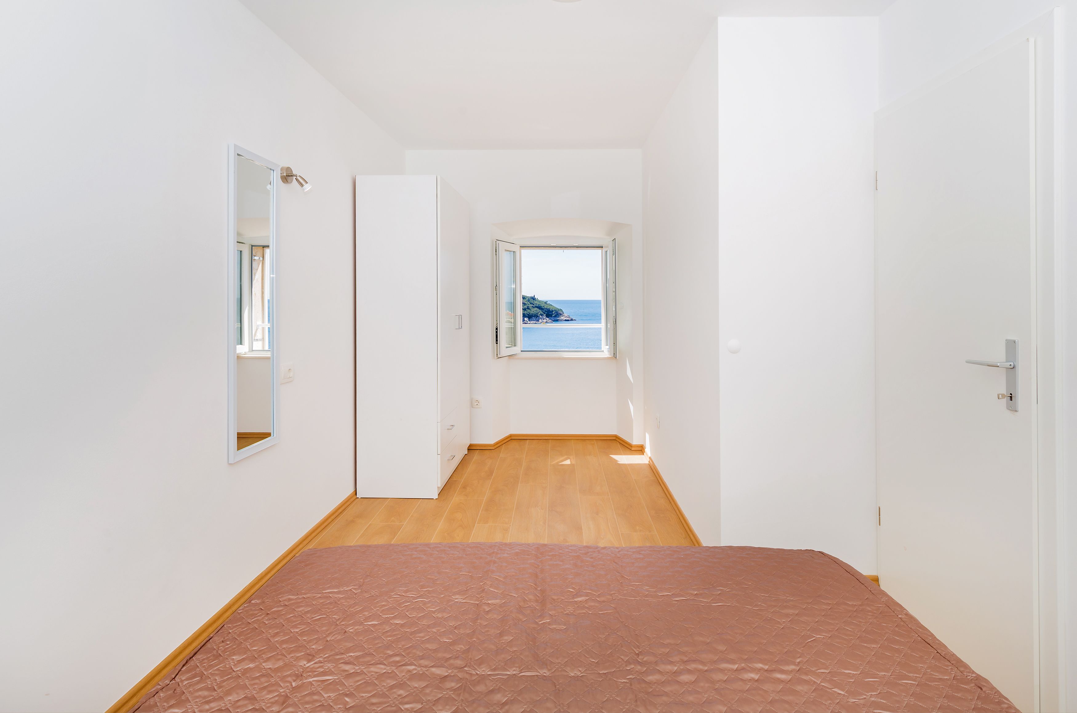 Un pasillo estrecho con una cama al final, una ventana con vista al mar y una puerta a la derecha.