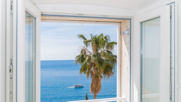 A room with a large window overlooking a beach with palm trees and the sea. - Apartment View (Dubrovnik)