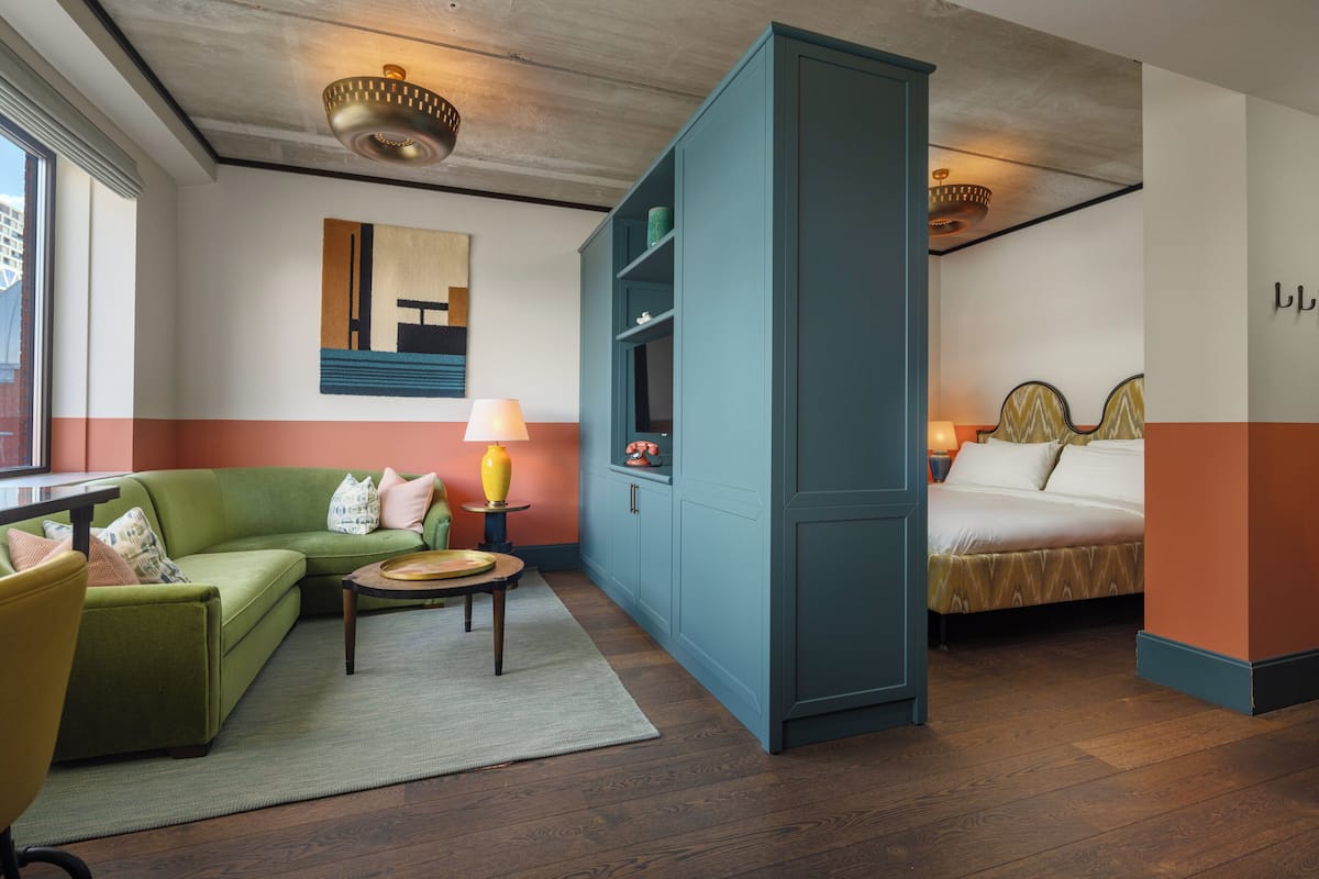 A modern hotel room with a green sofa, a bed, a blue wardrobe, and a small table with a lamp.