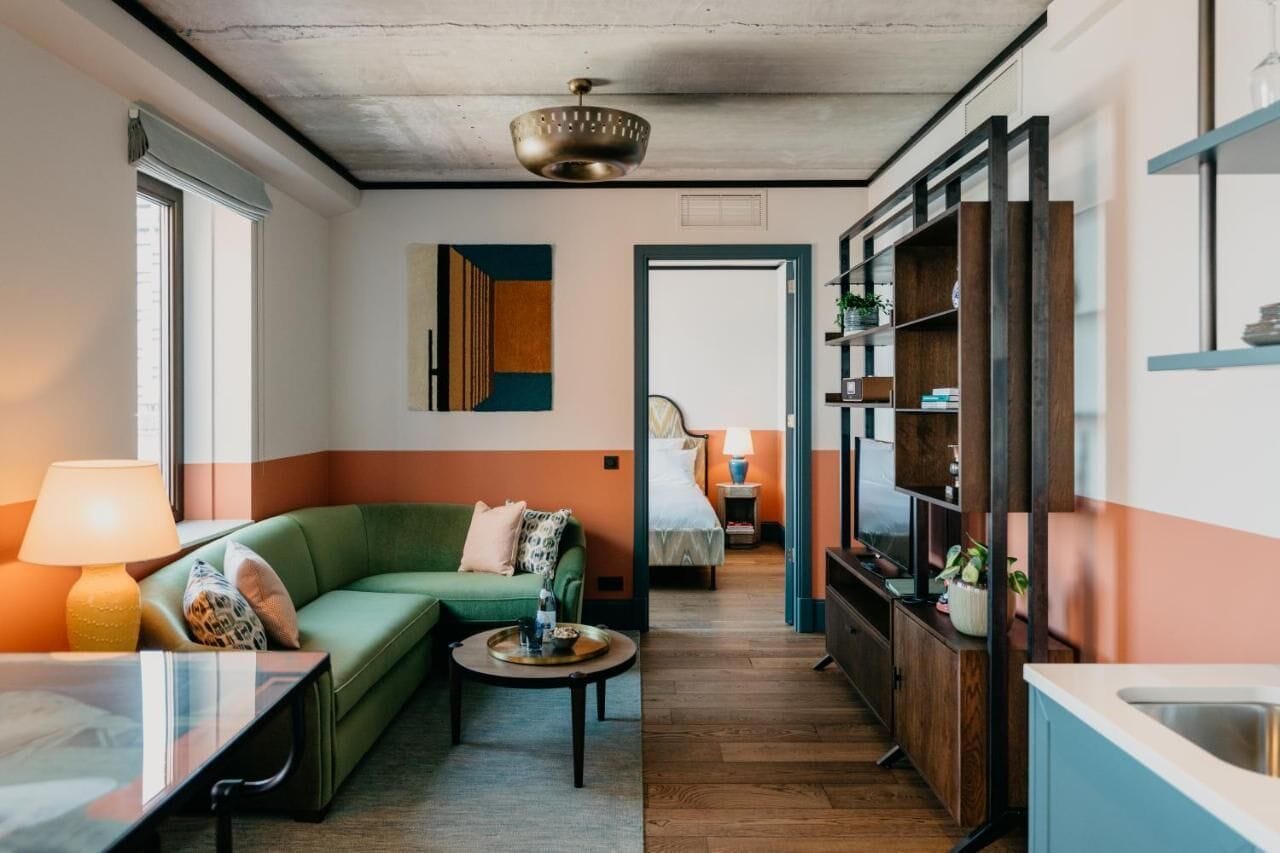 A modern living room with a green sofa, a coffee table, and a TV. There is a kitchen area with wooden cabinets and a sink.