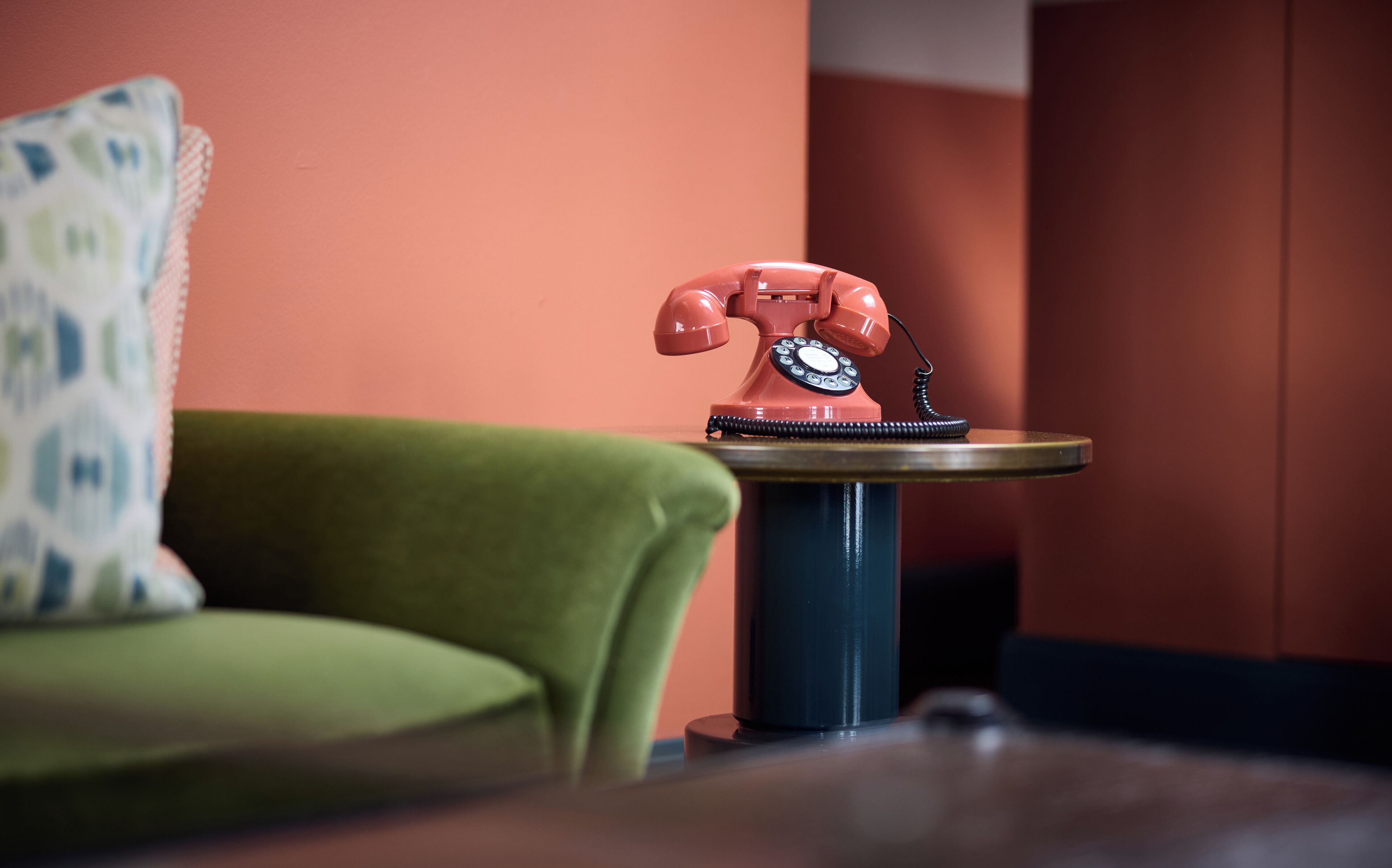 Een rode vintage telefoon met draaischijf op een klein rond tafeltje in een kamer met een groene bank en een kussen met dessin.