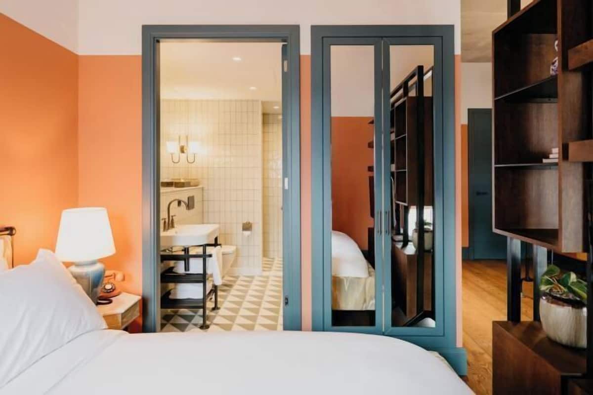 A hotel room with a bed, a nightstand, a lamp, a wardrobe, and a bathroom visible through an open door.