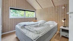 Une chambre avec un plafond en pente, des murs en bois, une grande fenêtre, un lit recouvert d’une courtepointe, une table de chevet avec une lampe et une petite commode.