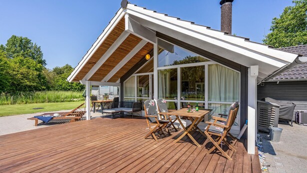 Un espace extérieur abrité, doté d’une terrasse en bois, d’une table avec des chaises et d’une grande fenêtre donnant sur un paysage verdoyant.