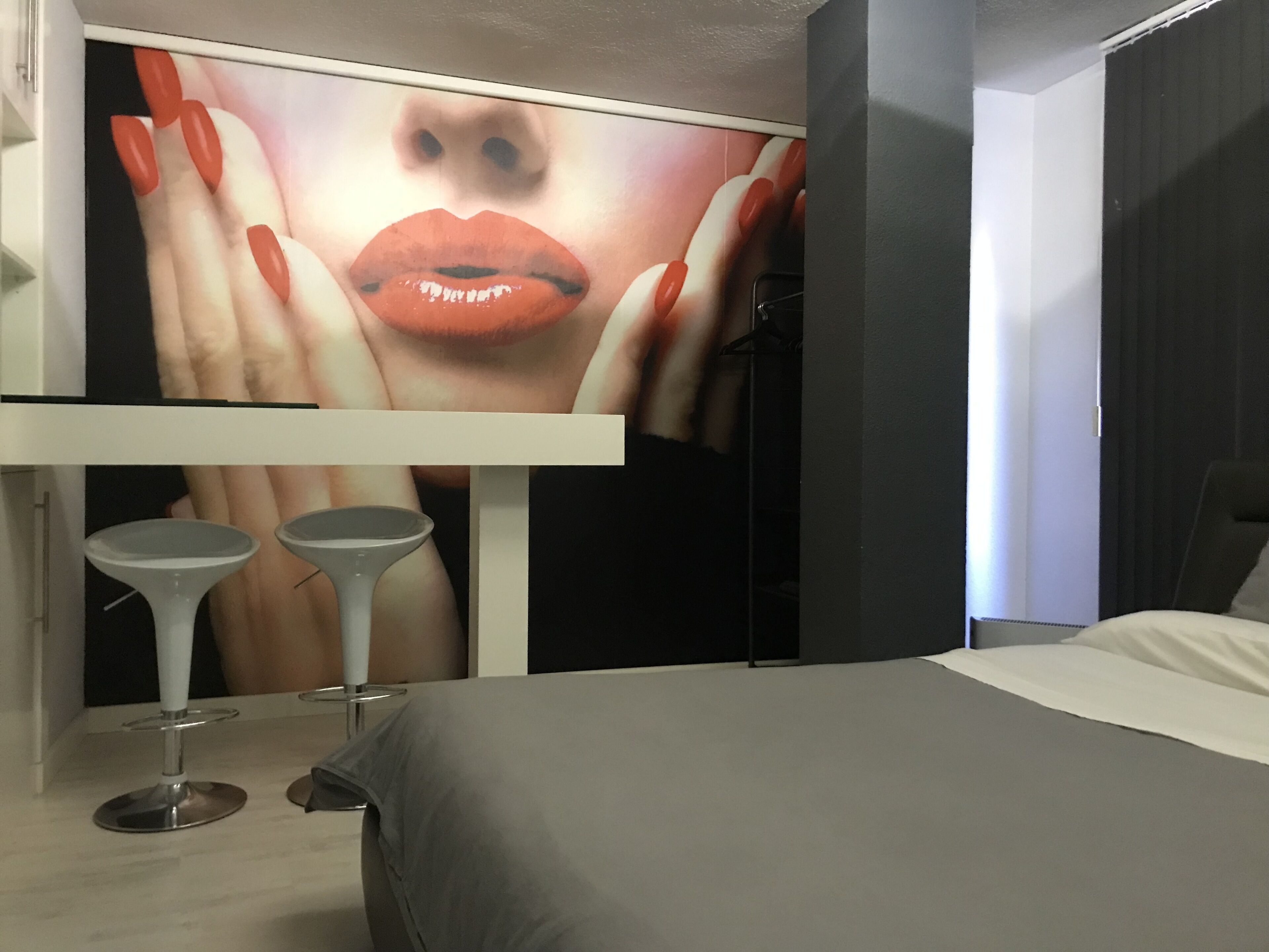 Ein modernes Hotelzimmer mit einem großen Bett, einem kleinen Tisch mit zwei Hocker und einem an der Wand befestigten Fernseher, der ein Bild von Lippen zeigt.