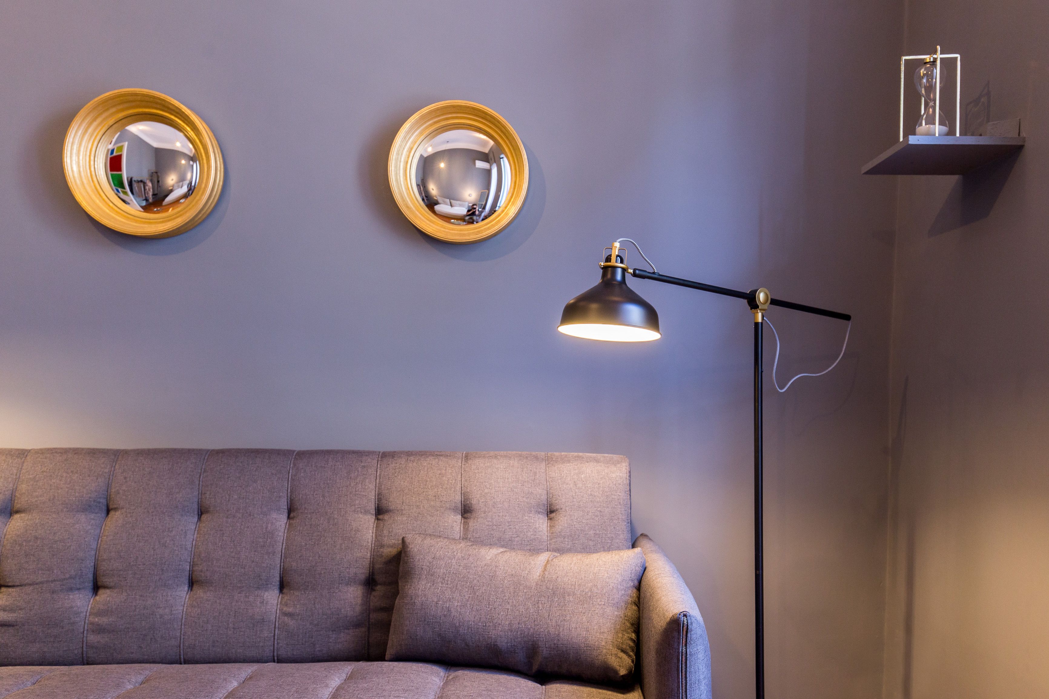A room with a tufted sofa, two wall-mounted mirrors, a floor lamp, and a shelf with a decorative item.