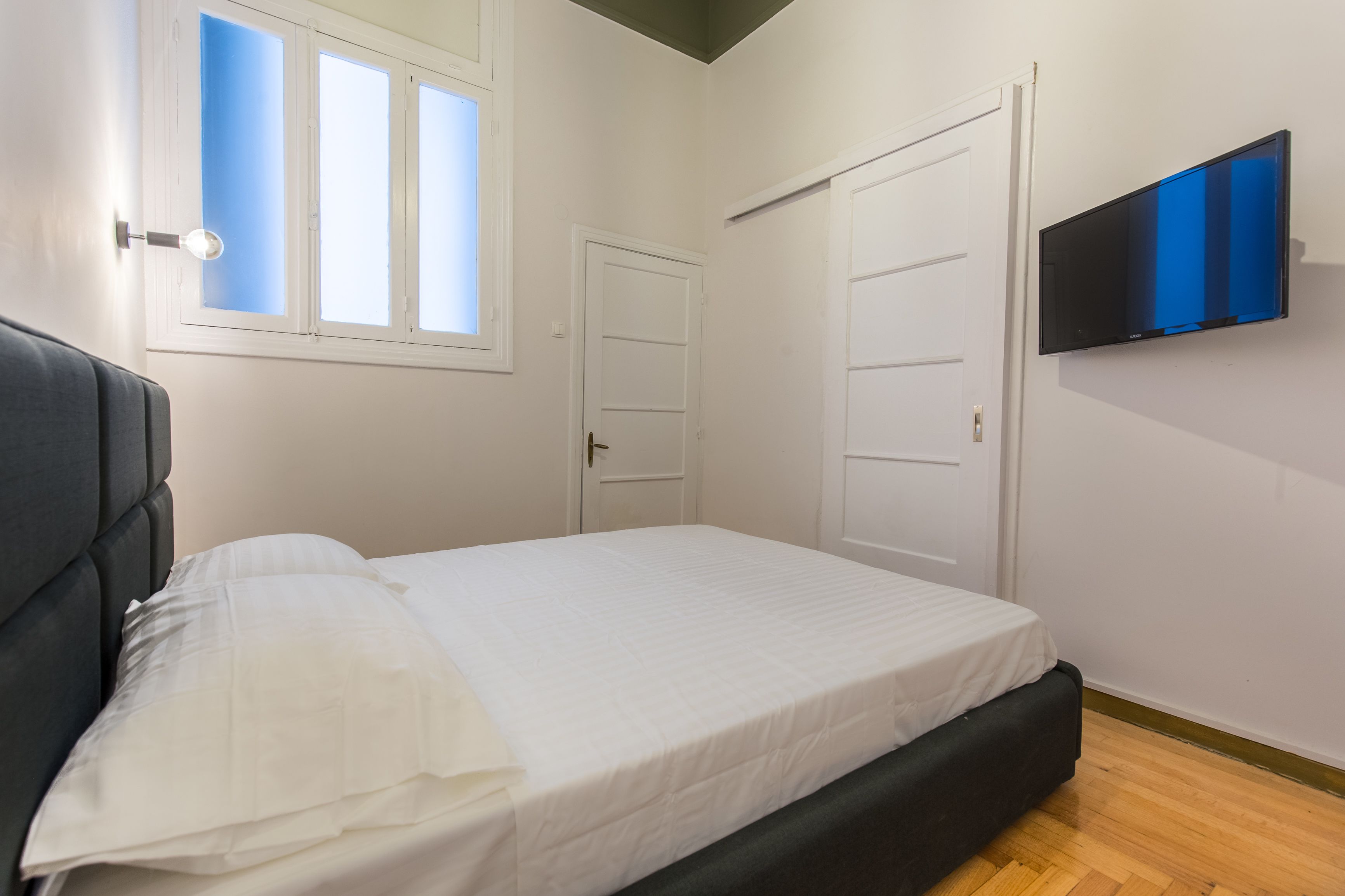 A bedroom with a bed, a TV mounted on the wall, and a window with blinds.