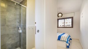 A compact room with a glass-enclosed shower, a bed with a striped bedspread, and a window with barred glass.