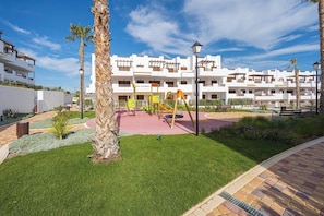 A playground with swings and slides, surrounded by palm trees and a building with balconies.