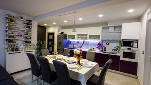 A modern kitchen with white and purple cabinets, a dining table set for four, and a wine rack on the left. - Villa Hedonist (Seget)