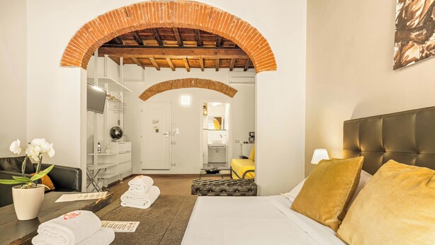 A hotel room with a bed, a sitting area with a chair and a table, a mirror, a TV, and a bathroom visible through an archway. - Canto dei Servi (Florence)
