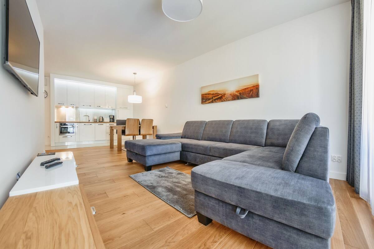 Een moderne woonkamer met een grijze modulaire bank, een houten salontafel en een keuken op de achtergrond.