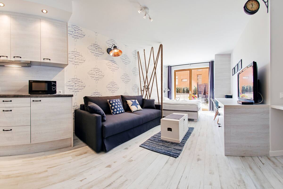 Een moderne woonkamer met een donkere bank, een klein witte salontafel, en een keukenhoek met een magnetron.