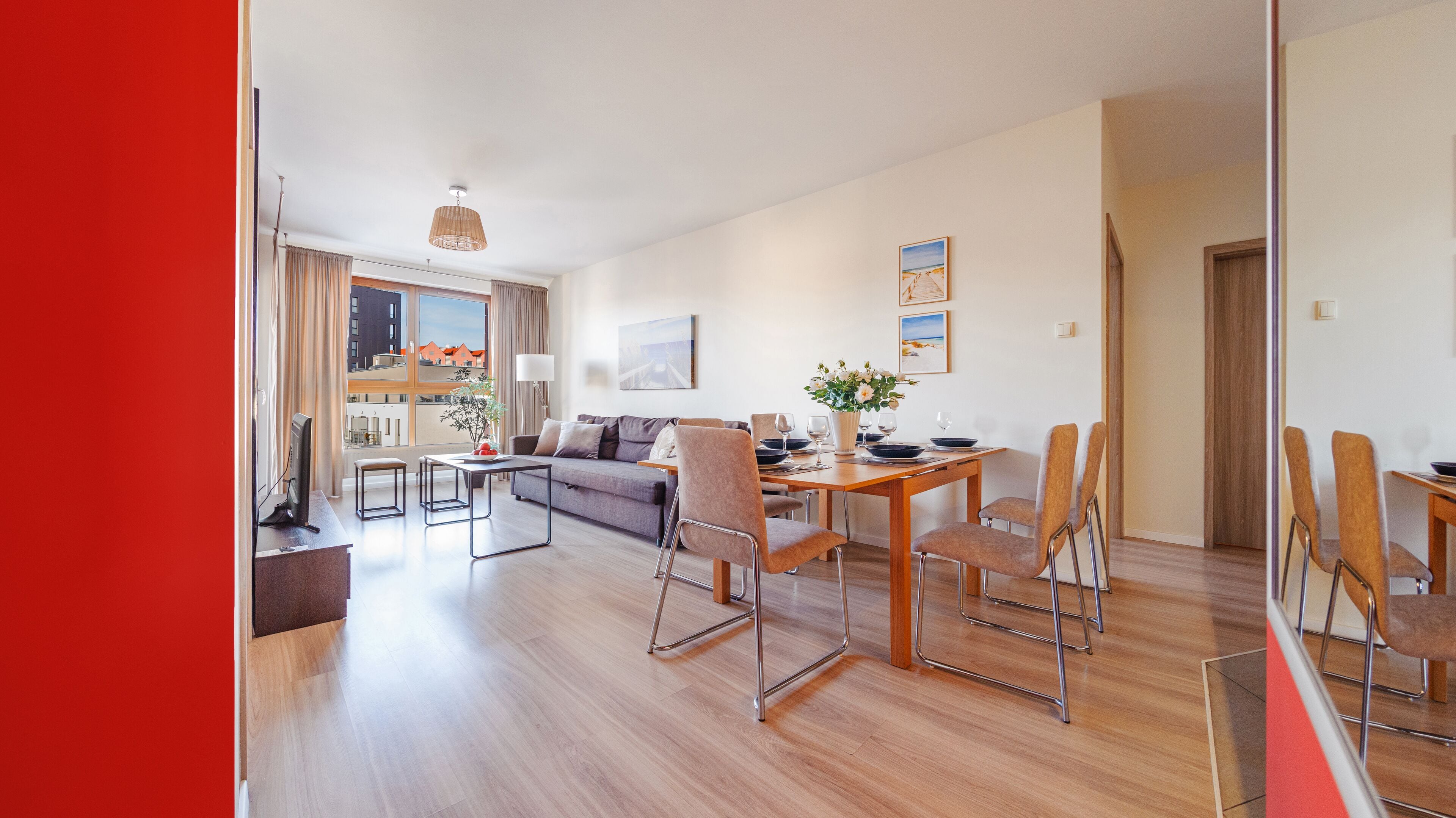 Una sala moderna con sofá, mesa de comedor y sillas. Hay una ventana con vistas a edificios y una puerta que da a otra habitación.