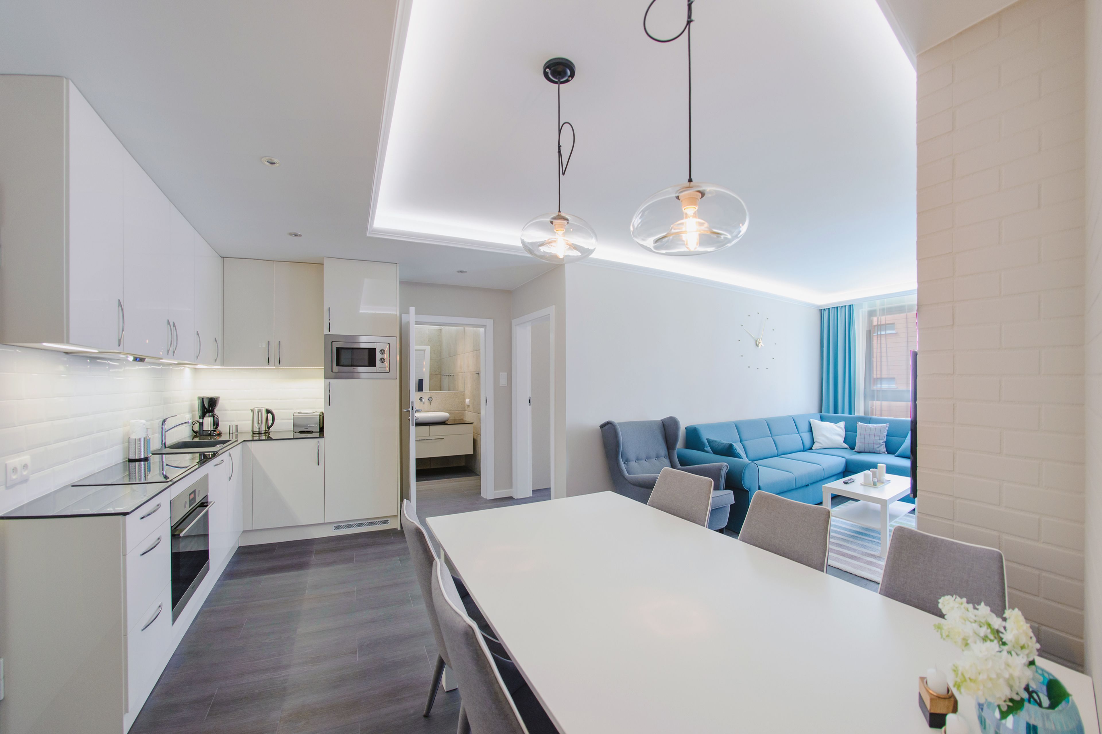 Una cocina moderna con armarios blancos, un comedor con mesa blanca y un sofá azul en la sala de estar.