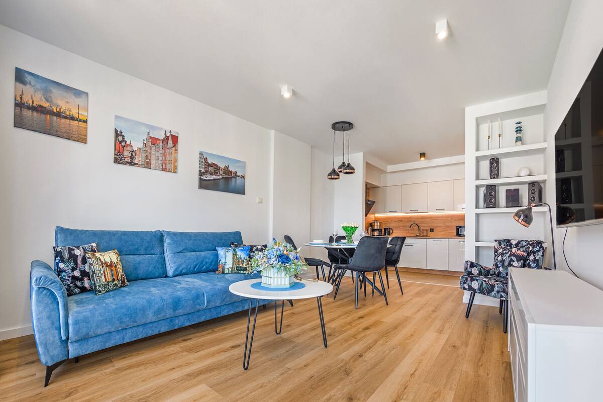 Een moderne woonkamer met een blauwe bank, een kleine ronde tafel en een eethoek met stoelen. Er zijn keukenkastjes en een televisie die aan de muur is bevestigd.