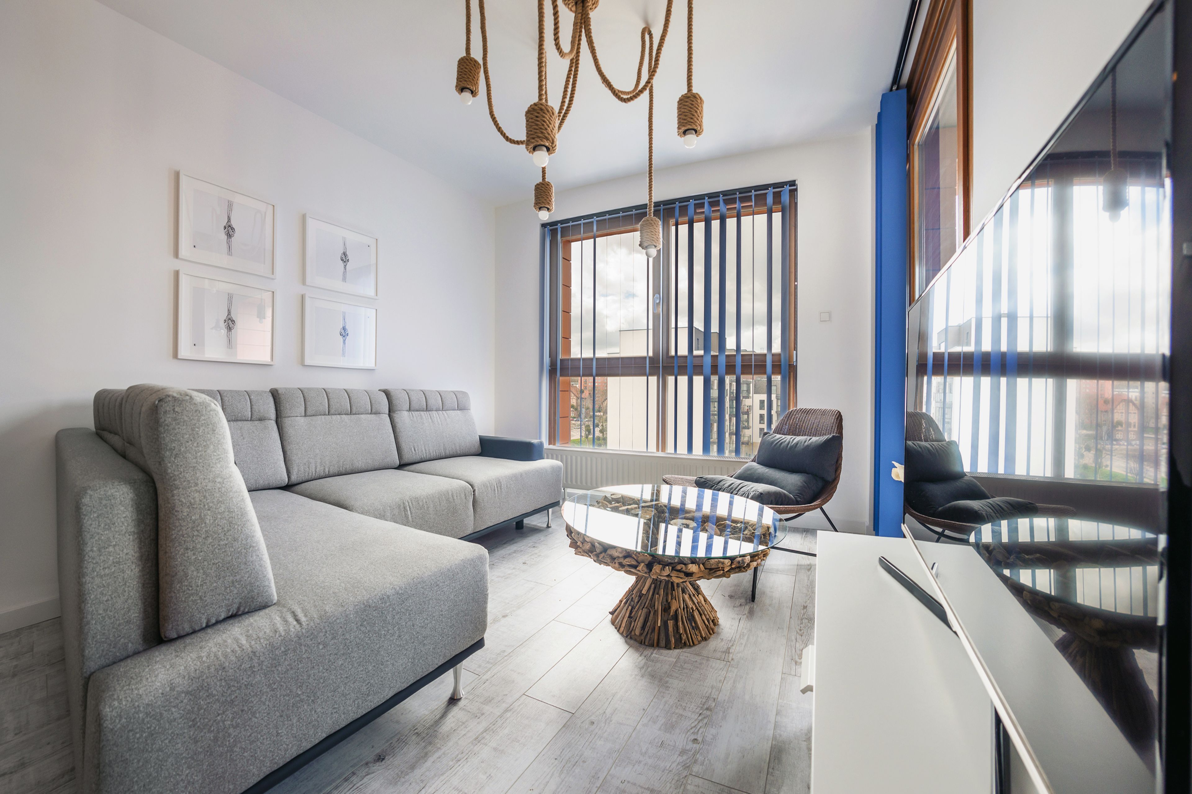 Un salon moderne avec un canapé d’angle gris, une table basse en verre et un téléviseur posé sur un meuble blanc. Des tableaux encadrés ornent le mur, et une grande fenêtre est équipée de stores.