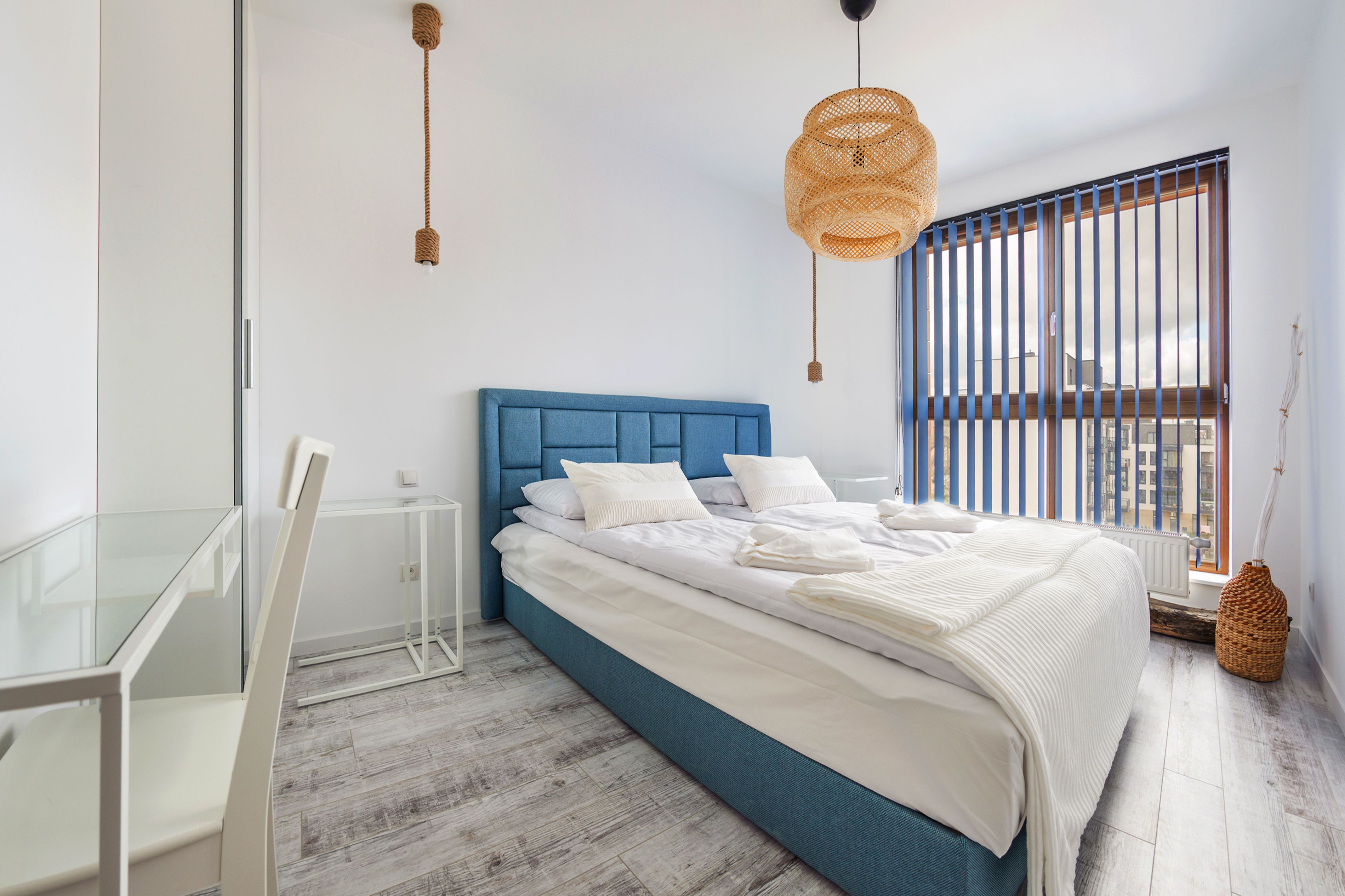 A bedroom with a blue headboard, white bedding, a glass desk, and a large window with vertical blinds.