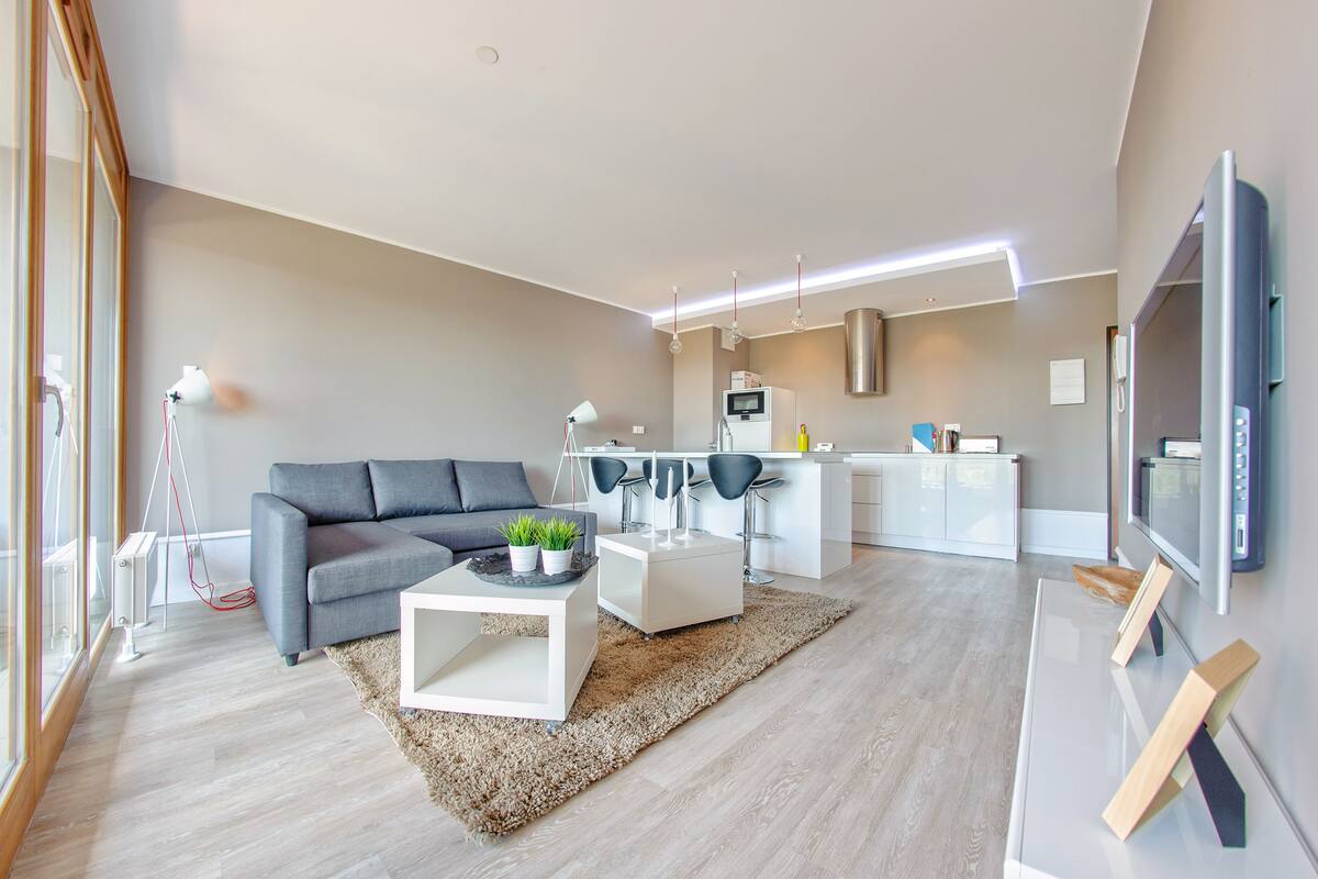 Een moderne woonkamer met een grijze bank, een salontafel en een televisie. Er is een keuken met een witte koelkast en een magnetron.