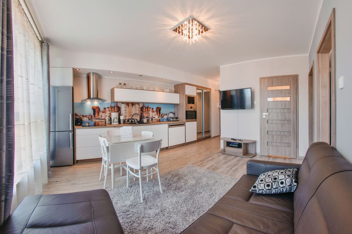 Een moderne woonkamer met een bruine bank, een witte eettafel en een keuken op de achtergrond.
