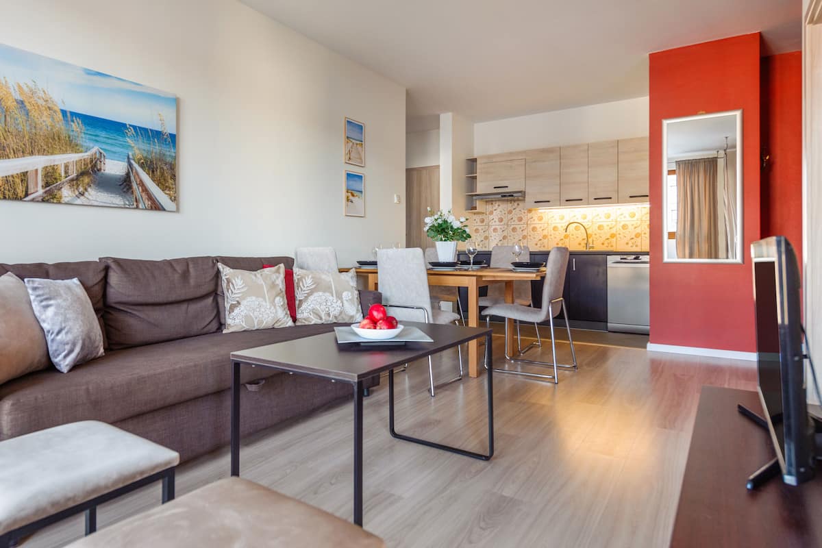 Een moderne woonkamer met een bank, salontafel en een eethoek. Achtergrond is een keuken met houten kastjes en een rode accentmuur.