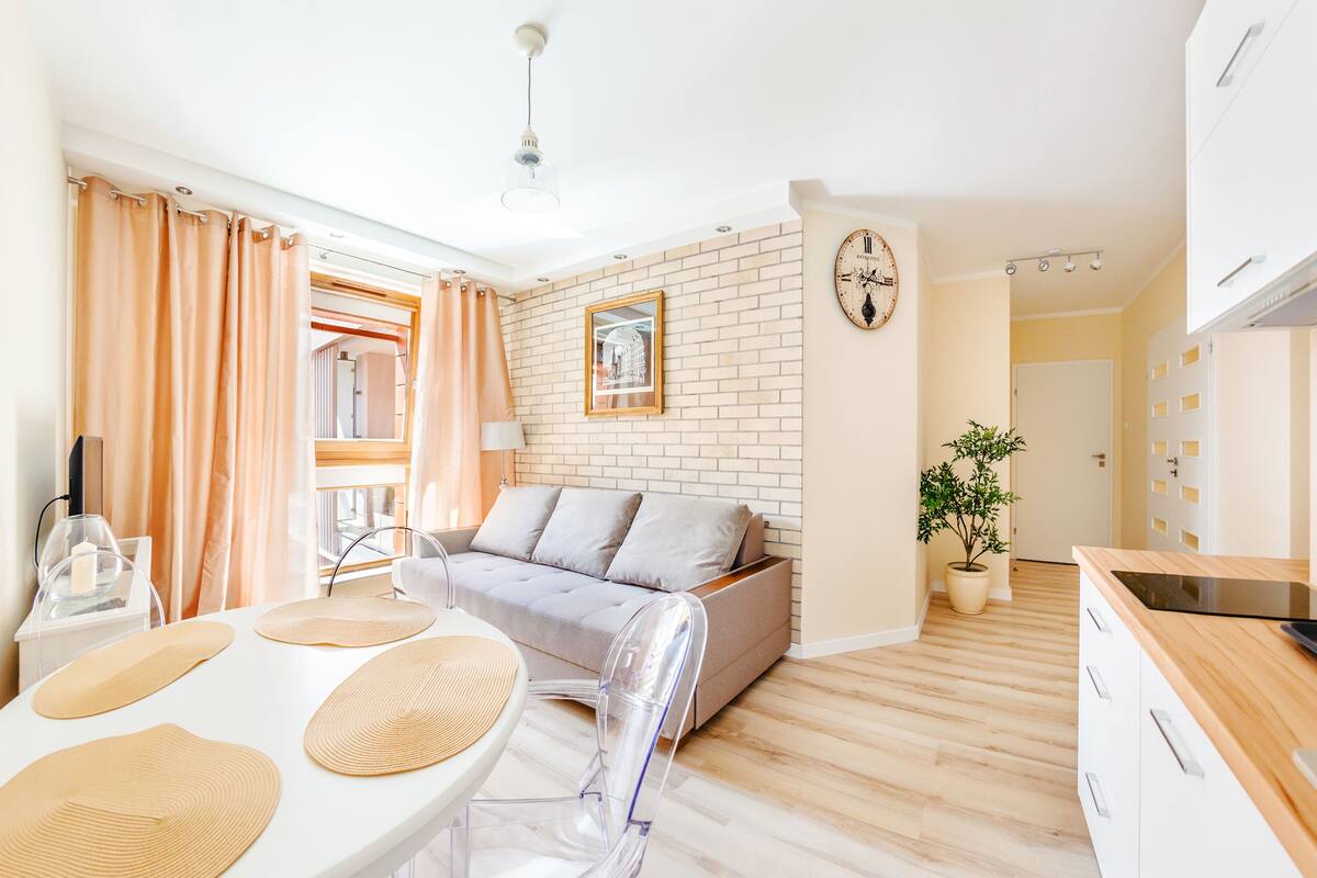Een moderne woonkamer met een bank, een eettafel met transparante stoelen, een potplant en een keuken met aanrecht en kastjes.