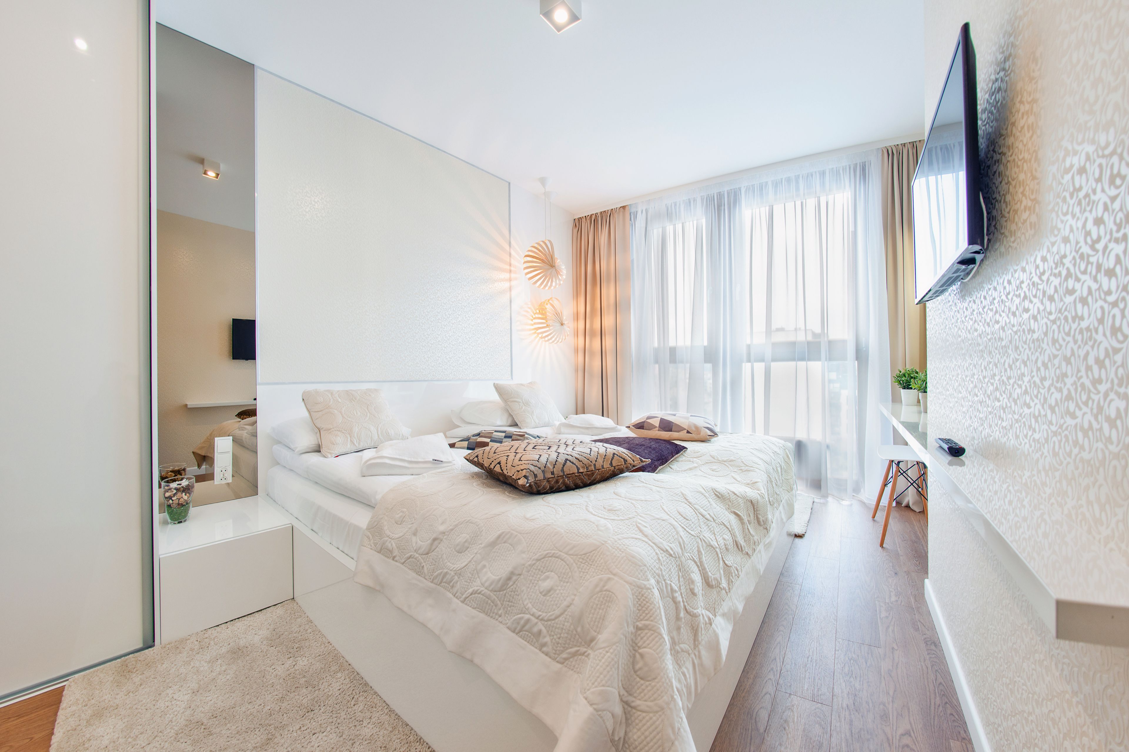 A modern bedroom with a large bed, a flat-screen TV mounted on the wall, and a large window with sheer curtains.