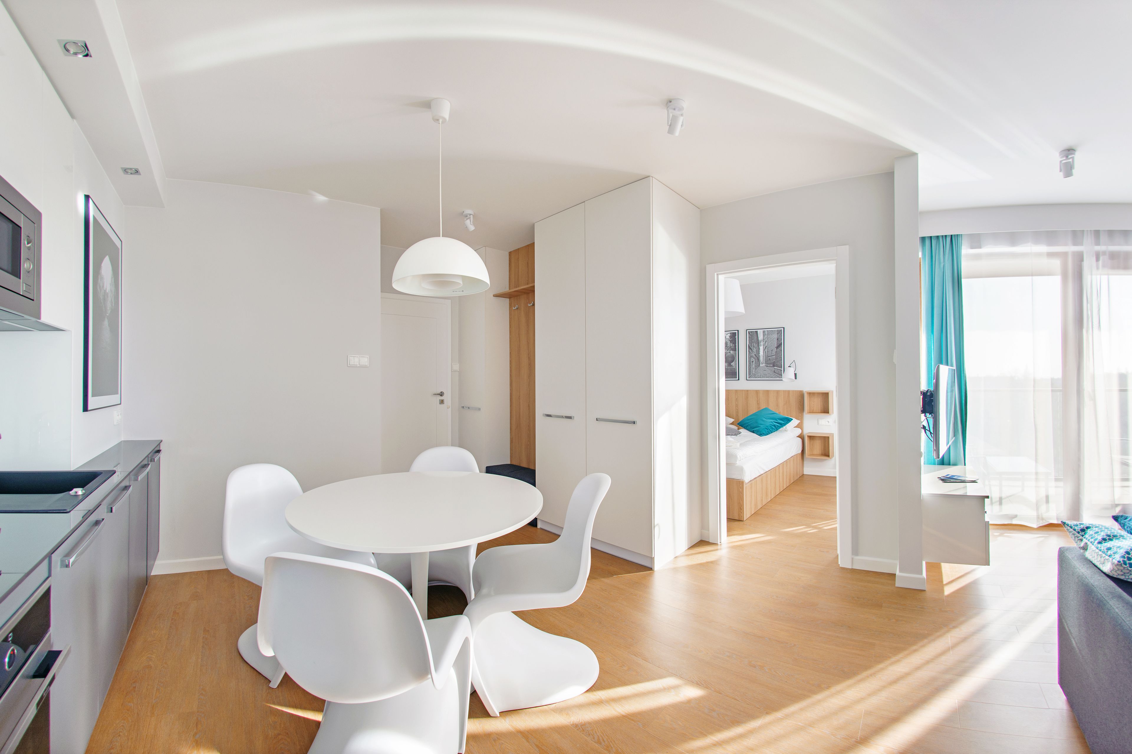 Una habitación moderna y luminosa con una mesa de comedor redonda y sillas blancas, una zona de cocina con microondas y vistas a un dormitorio con cama y escritorio.
