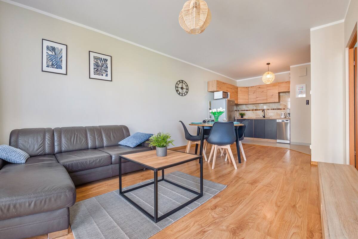Een moderne woonkamer met een leren bank, een eettafel met stoelen, een keukenhoek en een groot raam met gordijnen.