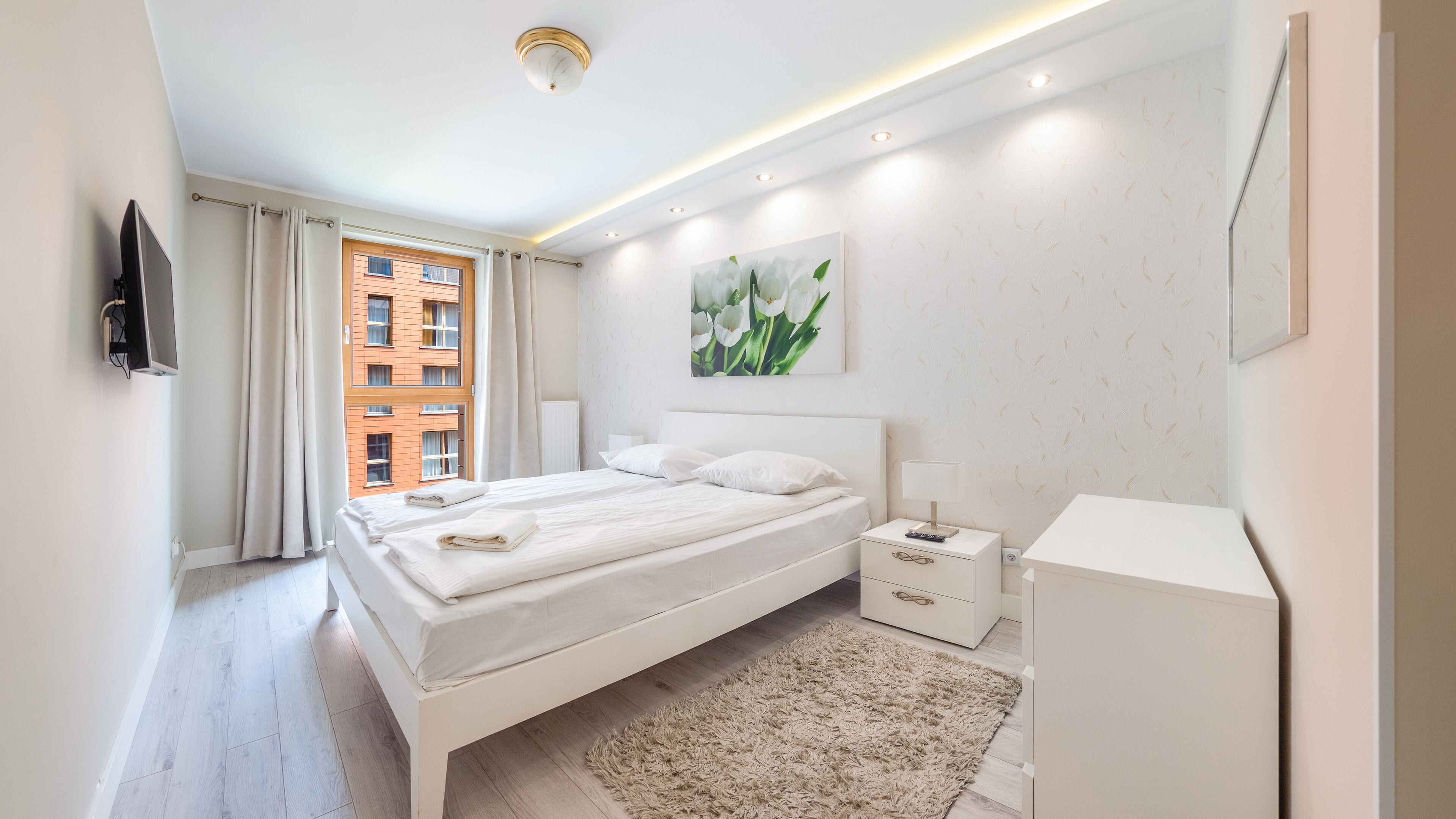 Une chambre moderne avec un grand lit, des tables de chevet, une commode, un téléviseur et une fenêtre avec des rideaux.