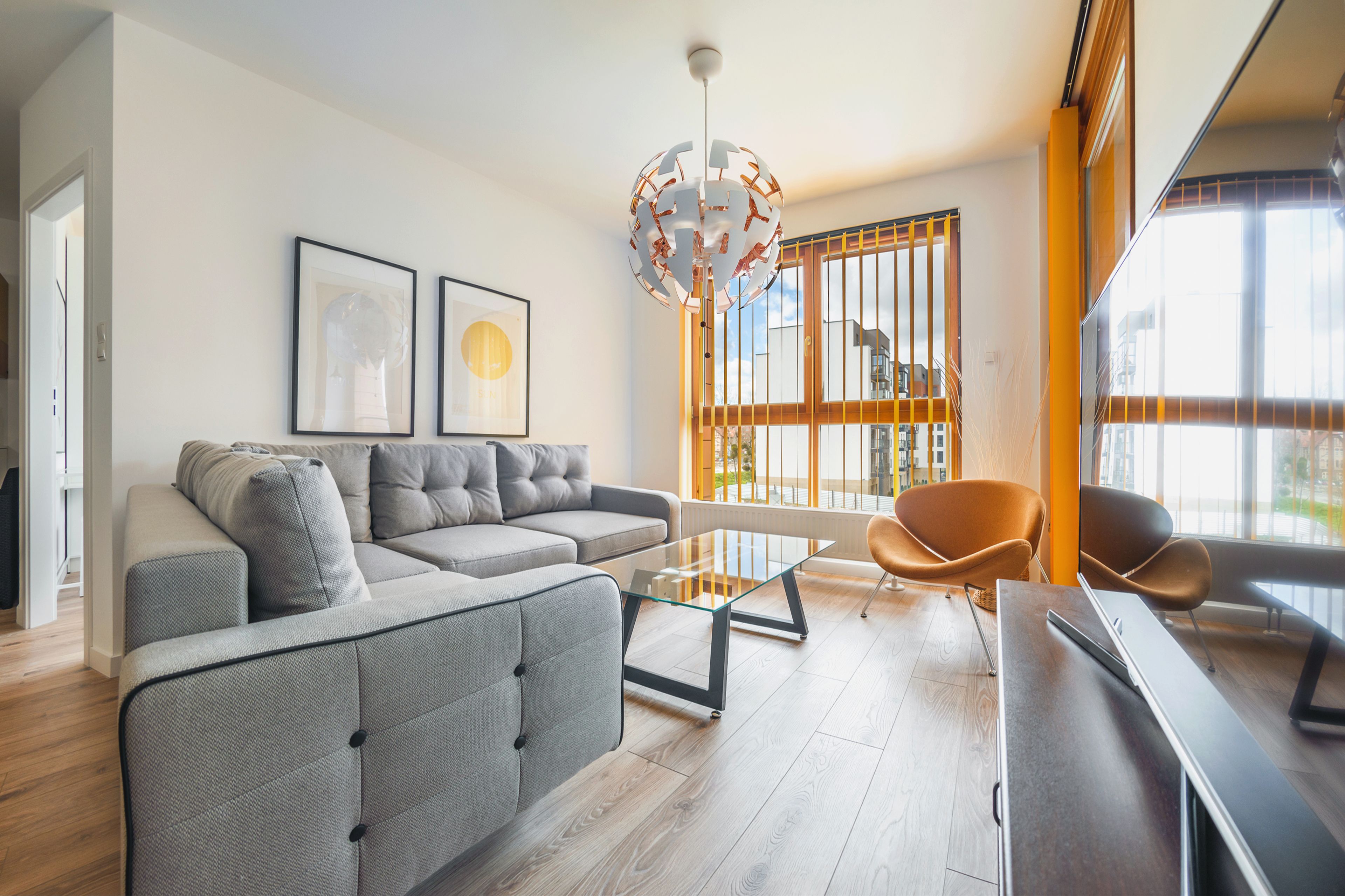 Una sala de estar moderna con un sofá gris, una mesa de centro de vidrio, sillas naranjas y un ventanal con vistas a edificios.