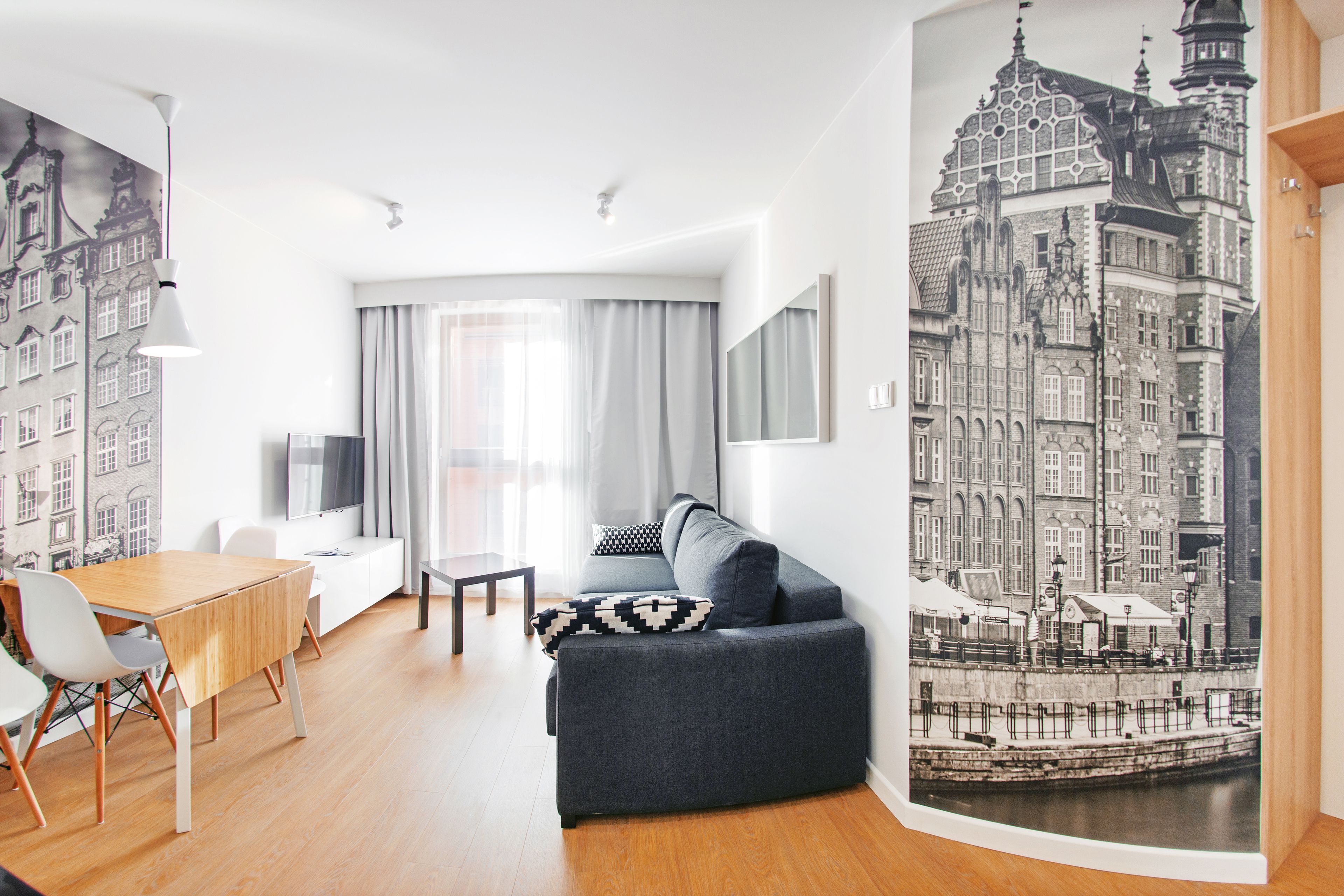 Een moderne woonkamer met een bank, eettafel en stoelen. Er is een groot raam met gordijnen, een flatscreen-tv en een kunstwerk op de muur, waarop een stadsgezicht te zien is.