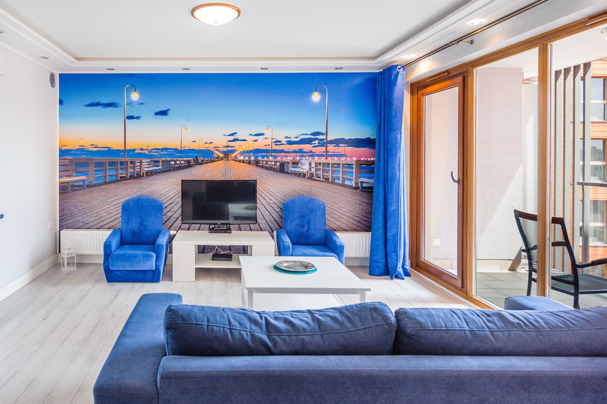 Een moderne woonkamer met een grote televisie, twee blauwe fauteuils, een witte salontafel en een bank.