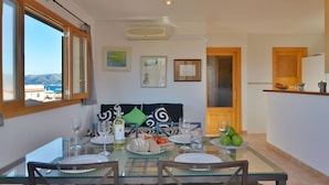 Espaço de refeições com mesa posta para dois, vista para o mar e cozinha com uma fruteira.