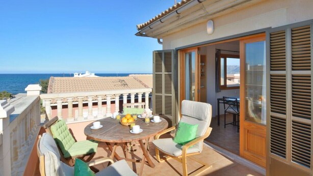 Terraço com mesa e cadeiras, vista para o mar e um edifício com telhado cerâmico.