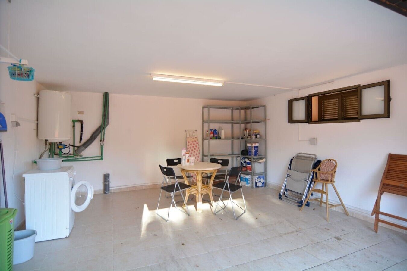 A room with a table, chairs, a shelf with items, and a window with blinds.