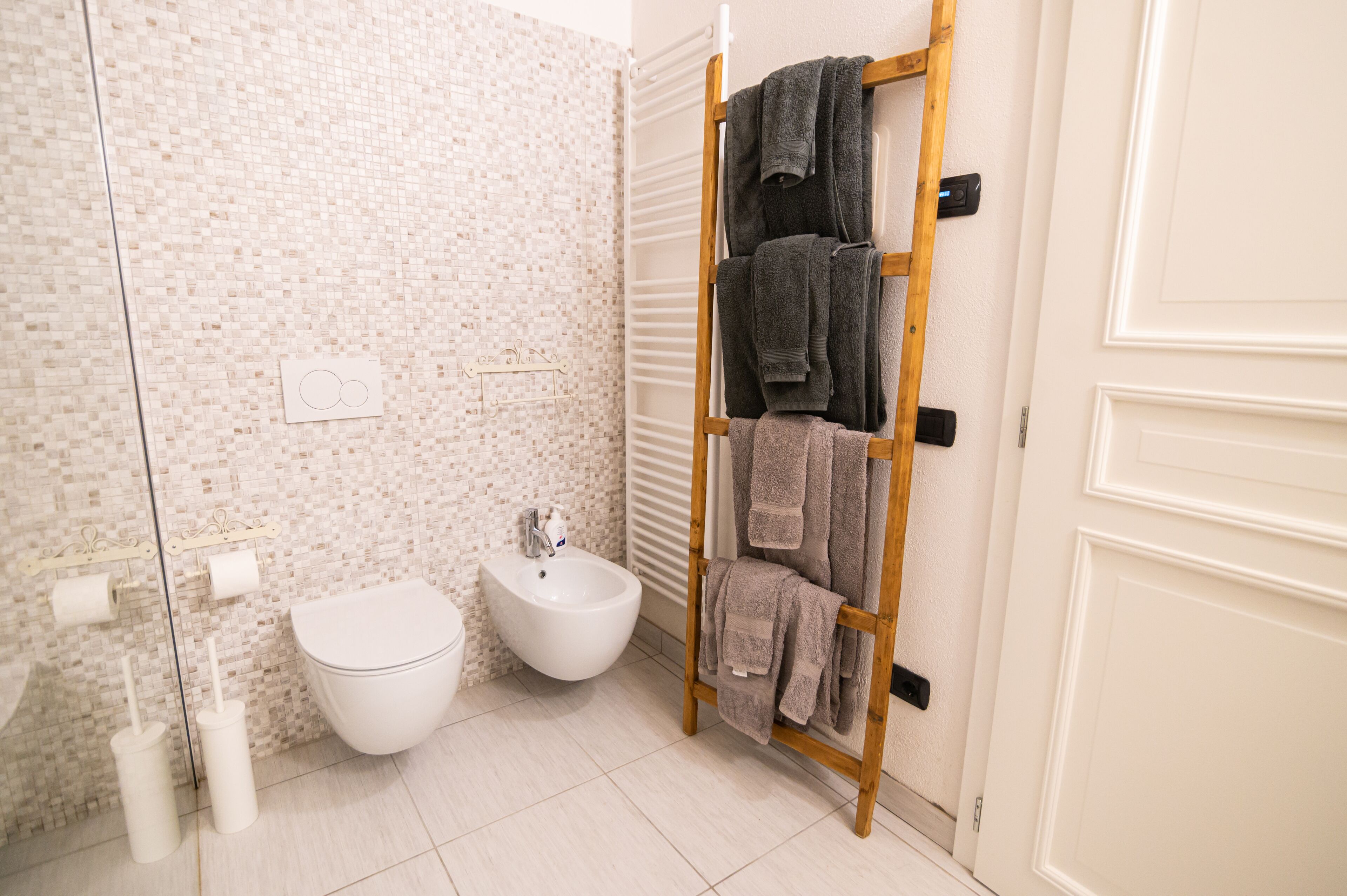 A bathroom with a bidet, two toilet paper holders, a towel rack with towels, and a white door.