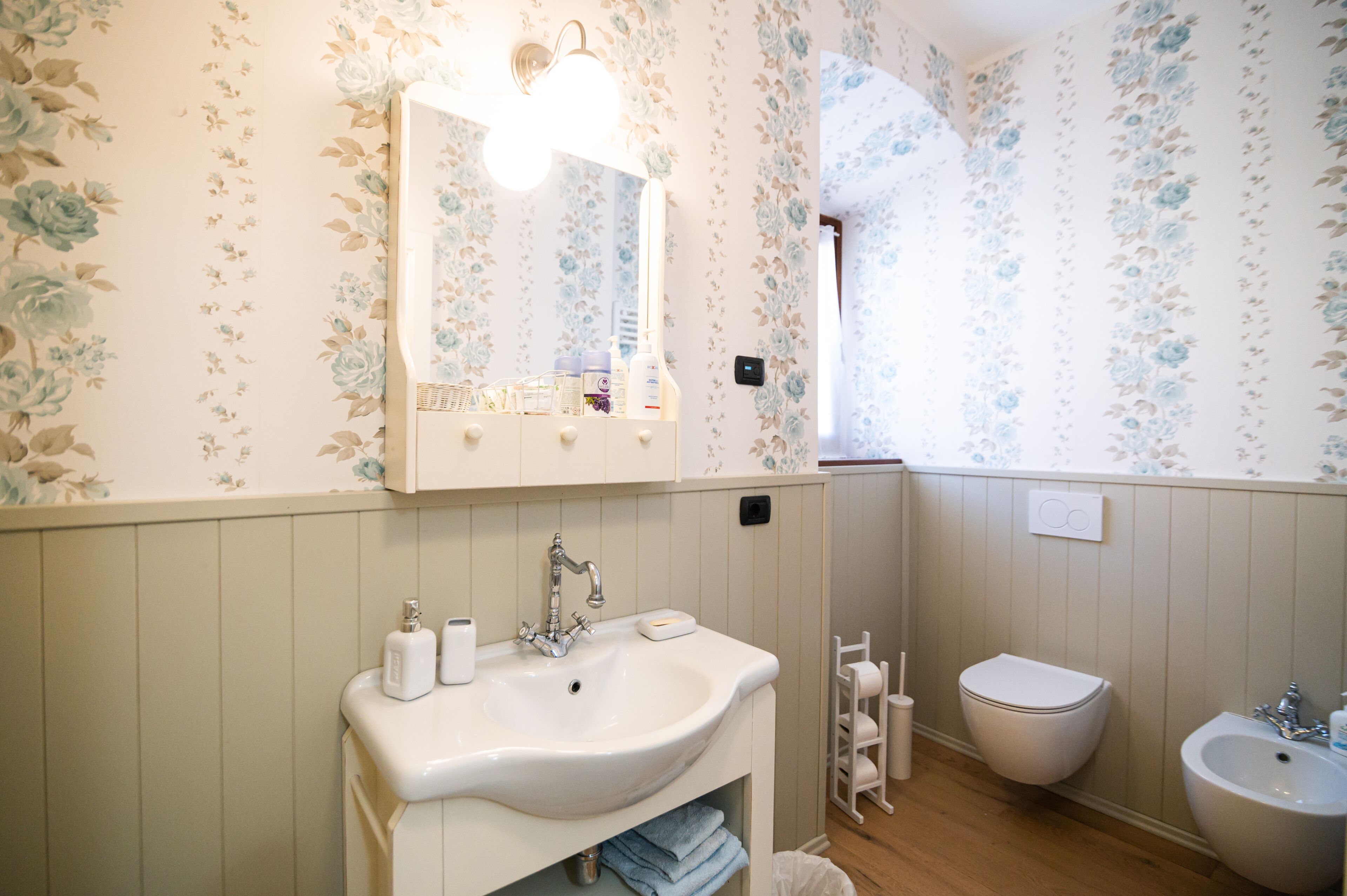 A bathroom with a white sink, mirror, and vanity, a toilet, and a bidet. The walls have floral wallpaper.