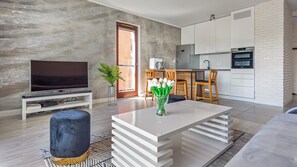 Ein modernes Wohnzimmer mit Flachbildfernseher, Couchtisch und Essbereich mit Stühlen. Im Hintergrund ist eine Küche zu sehen.