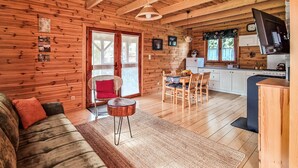 A cozy cabin interior with a wooden floor, a sofa with cushions, a dining table with chairs, a kitchen area with a stove and sink, and a television mounted on the wall.