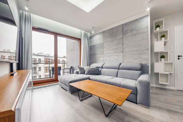 A modern living room with a grey sectional sofa, a wooden coffee table, and a large window with curtains.