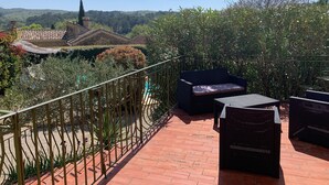 Terrassa amb moble de vimet, barana de metall i vista a una església i turons.