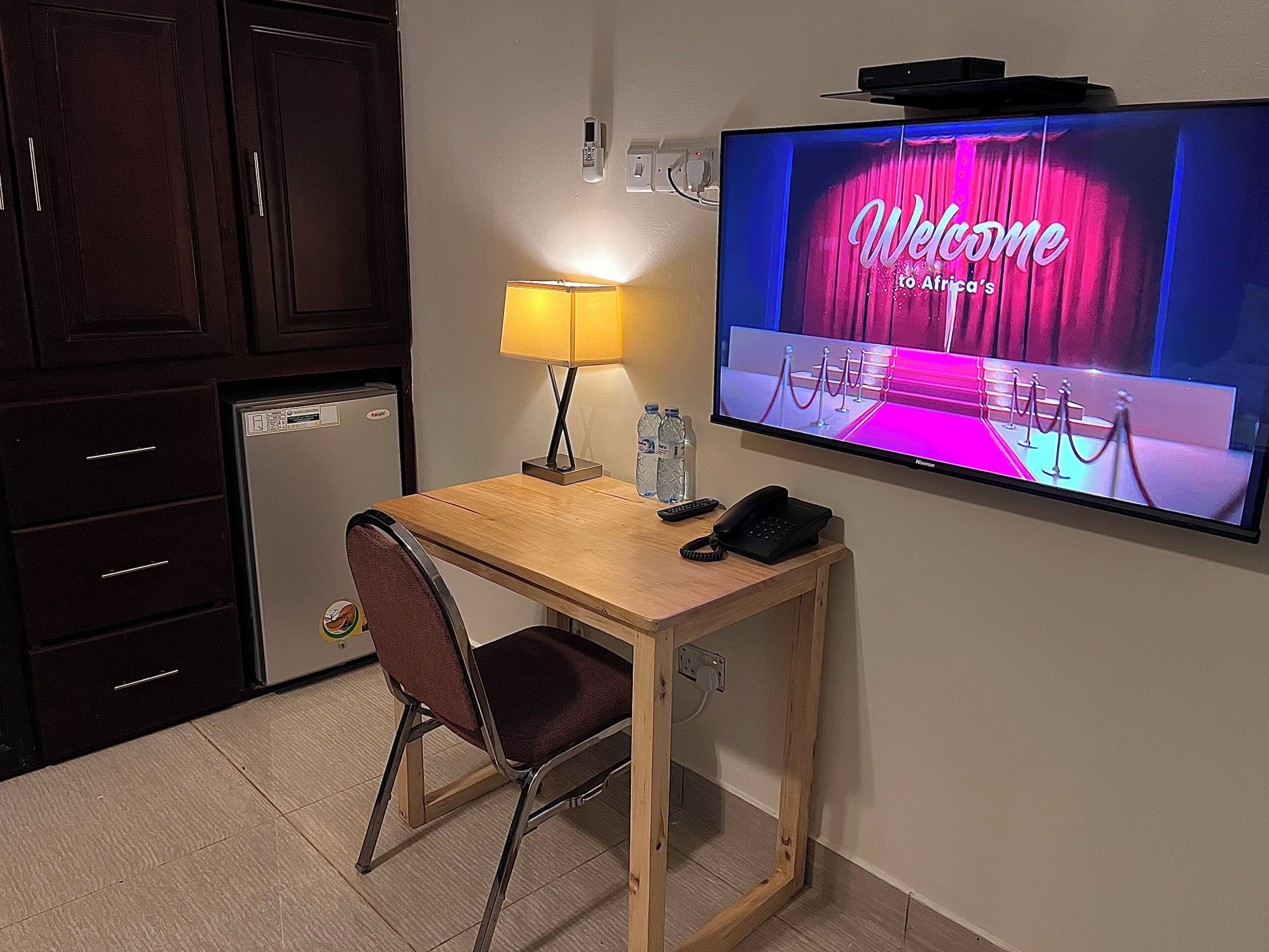A hotel room with a wooden desk, a chair, a television displaying a welcome screen, a lamp, a phone, and a minibar.