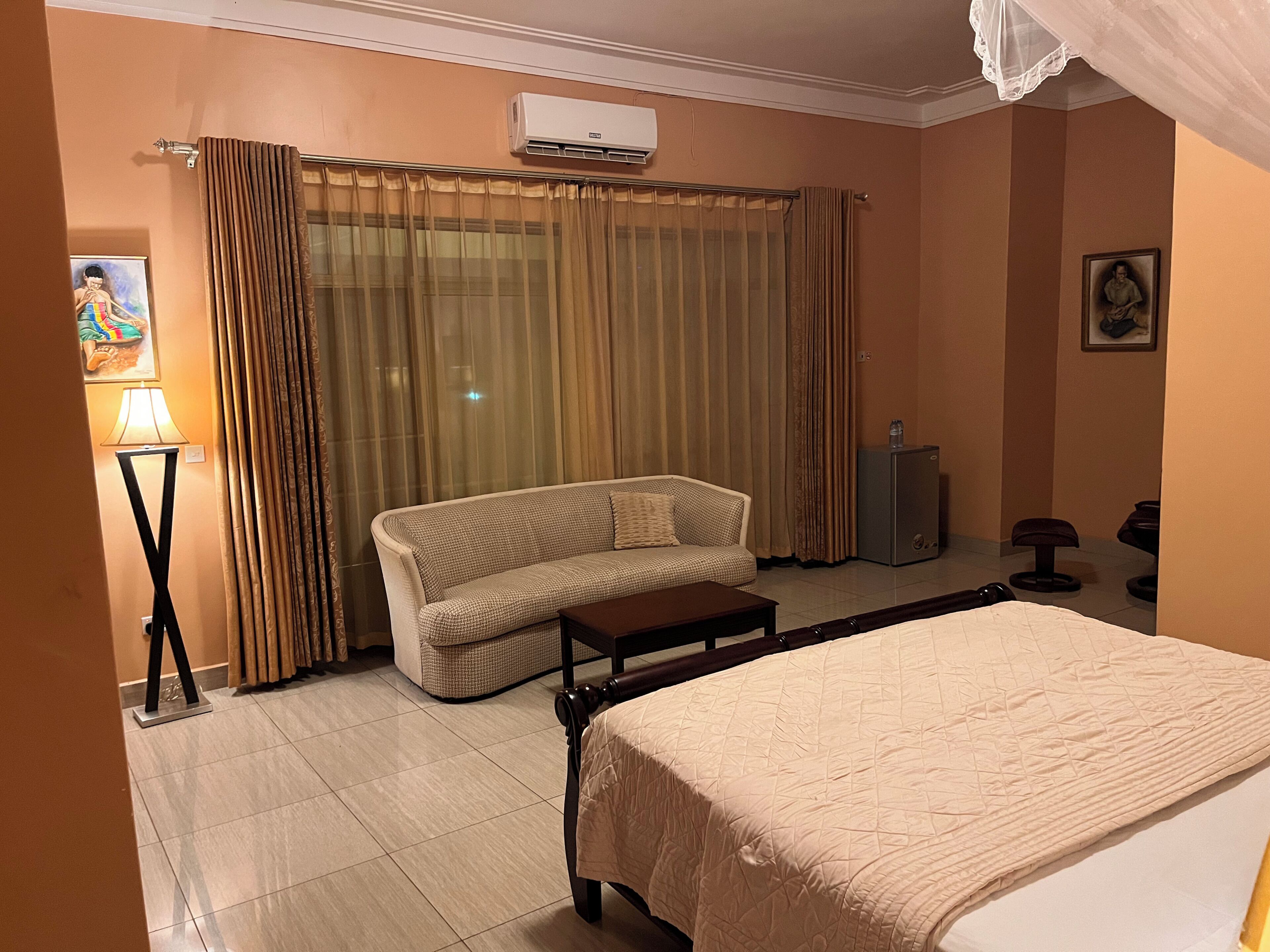 A hotel room with a bed, a sofa, a small table, an air conditioning unit, a lamp, curtains, and a picture on the wall.